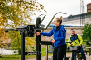 En glad kvinna står vid ett Omnigym utegym och greppar ett viktmagasin medan en man i gul tröja sitter i bakgrunden en höstdag.