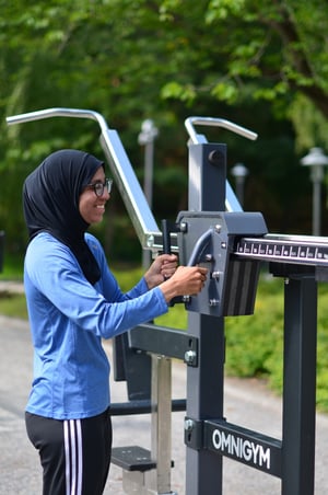 En leende kvinna i hijab justerar viktmagasinet på ett utegym i Trekantsparken och visar gymmets välkomnande miljö för alla.