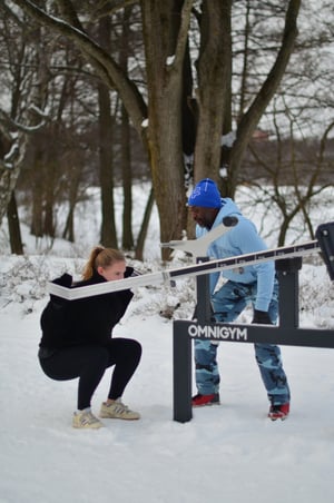 Winter scene at Trekantspark's outdoor gym: A man in blue coaches a woman performing squats, demonstrating the gym's year-round functionality and support.