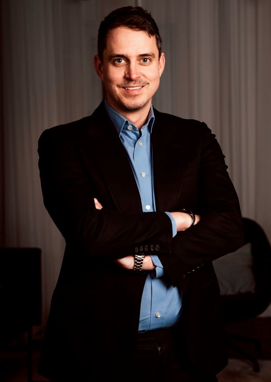 Mattias Svensson poserar med korsade armar och ett självsäkert leende, klädd i en svart kavaj över en ljusblå skjorta, som säljare av utegym hos Omnigym.