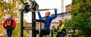 En kvinna tränar latsdrag och en man tränar tricepspress på ett utegym medan en annan man joggar förbi.