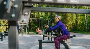Kvinna styrketränar på ett utegym, sittande i bicepsurl – i bakgrunden gör en annan kvinna crunches på en magbänk.