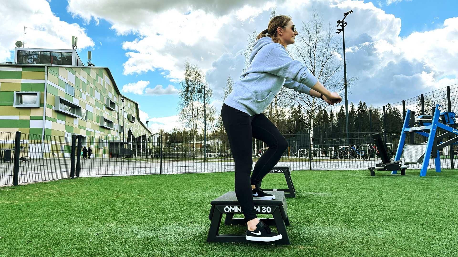 A woman doing an exercise standing on a 30 cm high plyo box in an outdoor gym.