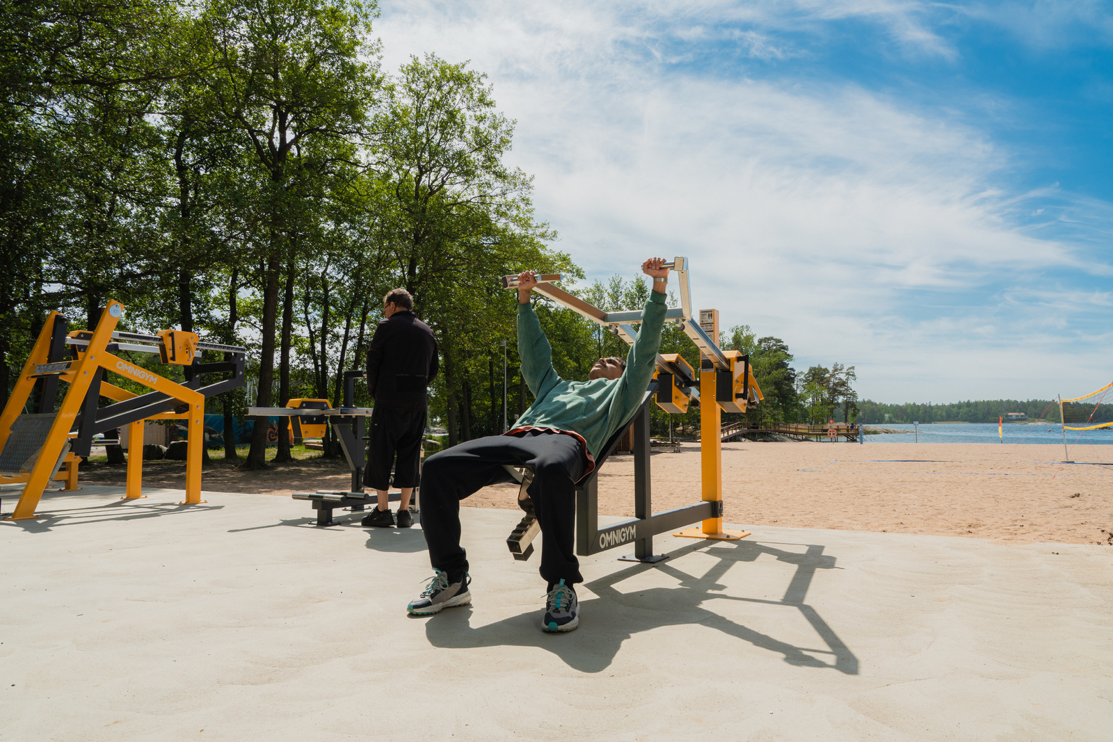 En man stryketränar lutande bänkpress på ett gult och svart utegym i stål vid en strand.