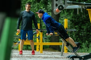 Two friends smiling while performing a Adjusted Back Extension exercise at an Omnigym outdoor gym.