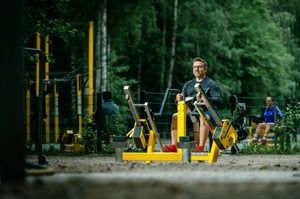 Mies hymyilee tehdessään soutulaite istuen -harjoitusta Omnigym-ulkokuntosalilla. Tämä voimaharjoittelu tapahtuu aurinkoisessa, avarassa ympäristössä, mikä tekee siitä täydellisen paikan kuntoilulle ulkona. Soutulaite istuen on tehokas harjoitus ylävartalon lihasten vahvistamiseen ja kestävyyden parantamiseen.