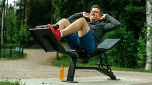 Man smiling while performing a adjustable abdominal bench exercise at an Omnigym outdoor gym, showcasing strength training in a sunny, nordic environment.