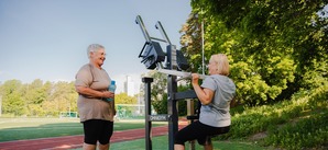 Två kvinnor tränar och pratar på ett utegym, en tränar latsdrag och en vilar. 