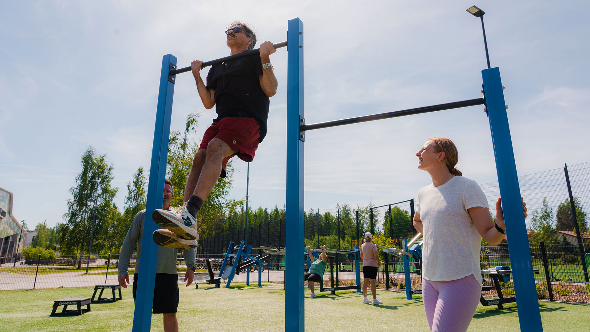 Homme et femme s’entraînant sur une aire de street workout en plein air, avec une barre de traction extérieure Omnigym. La femme réalise des relevés de genoux suspendus sur la barre, dans un espace de fitness extérieur moderne et équipé. Homme et femme s’entraînant sur une aire de street workout en plein air, avec une barre de traction extérieure Omnigym. La femme réalise des relevés de genoux suspendus sur la barre, dans un espace de fitness extérieur moderne et équipé.