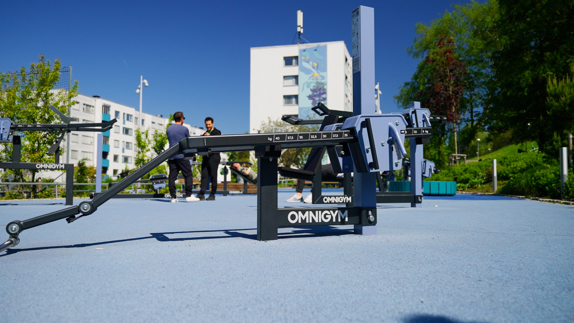 A close-up of a biceps curl at an Omnigym outdoor gym on a sunny day. Two men are standing in the background, talking